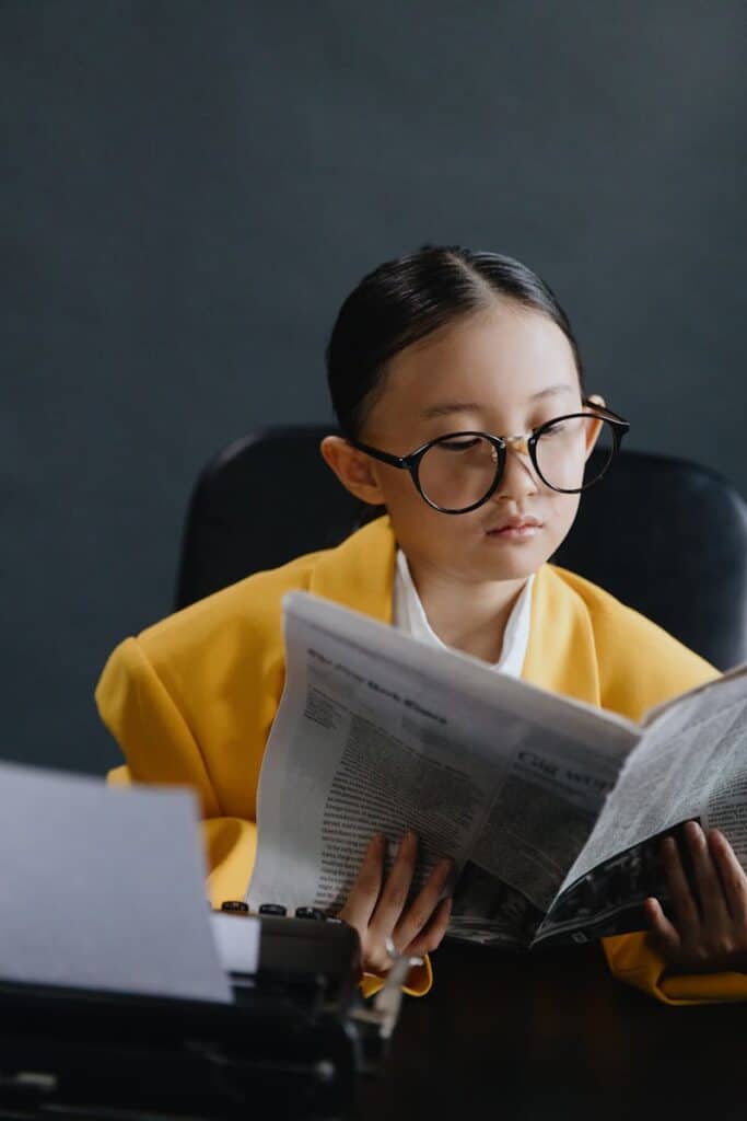 Child wearing a yellow suit and glasses, reading a newspaper indoors.